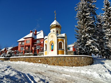 Chapel with the face of the saint on the wall and a dome sprinkled with snow. Standing near a large brick private house against the background of a cloudless winter sky.