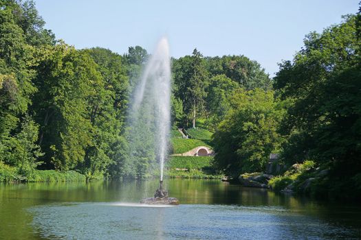 A large beautiful decorative fountain in the middle of a transparent lake against the background of a green park zone
