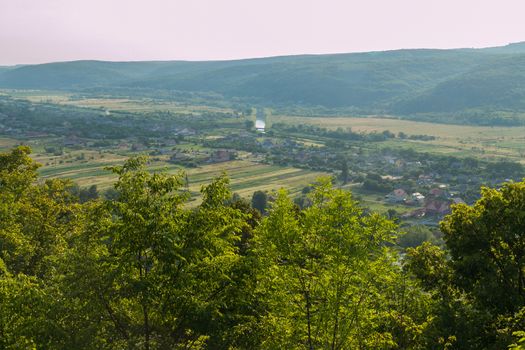 A picturesque view from a hill through the tops of deciduous trees to a cozy small village near a beautiful green mountain