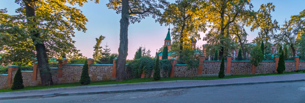 a panorama of tall trees growing next to the fence behind which the tops of domes can be seen