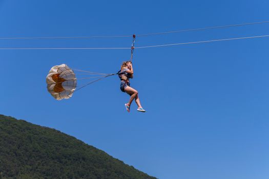 A woman riding along the cable clutching her hands with a stick with rollers tied for back insurance with a small parchute on the background of a green mountain slope and a blue sky.