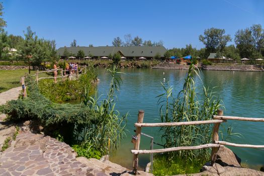 A large pond with paths lined with stone around it with walking people against the backdrop of a beautiful landscape around and a blue sky on them.