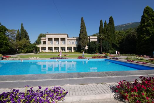 Transparent pool water with growing flowers along the edges located in the park next to a beautiful house against the blue sky and the prominent top of the mountain.