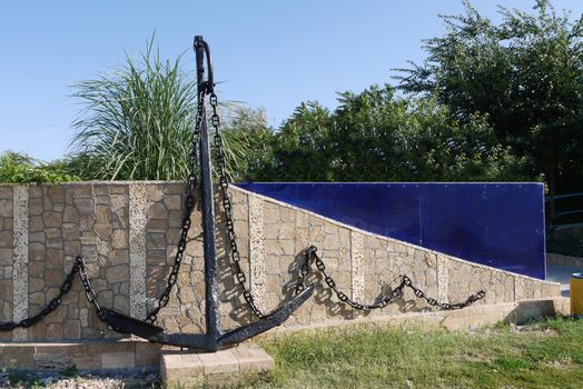 Marine anchor with a chain painted in black paint. Standing in the park leaned against a low wall made of stone.