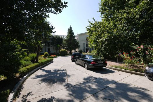 Several black cars parked on the sidewalk near the flowerbeds. In the background there is a white building