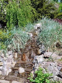 A beautiful artificial waterfall on flat stones flows between the plantings of ornamental grass