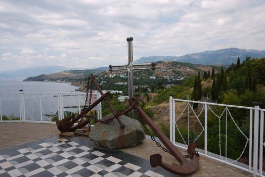 Rusted anchors with chains lying on a stone on a platform. Overlooking the coastal line of the sea and the house on the mountainside.