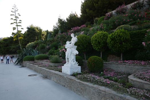 A beautiful interior in a park with flower beds with flowers standing several floors on a slope and green trees with spherical crowns growing there.