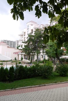 Beautiful view of a large white hotel with a swimming pool because of the greenery of bushes and trees growing on the flowerbed.