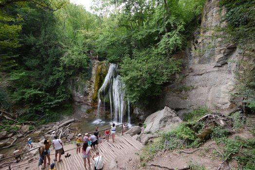 Tourists watch and photograph a beautiful mountain waterfall on a viewing platform near the rocky mountain