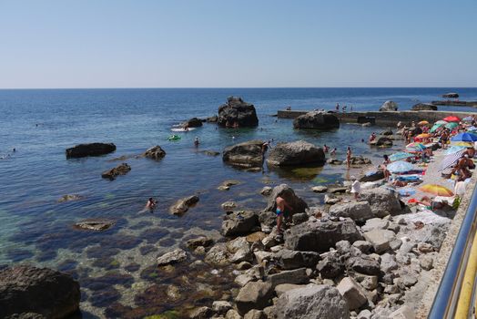 Tourists, resting on the beach next to the large stone boulders, going far into the sea