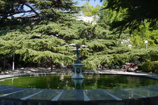 Recreation area with a huge fountain with a stone sculpture in the center against the background of green coniferous trees