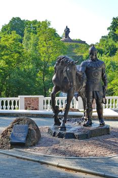 A statue of a knight in a helmet holding a horse against the background of green trees on a slope and a monument to a warrior on top with a sword lowered down.