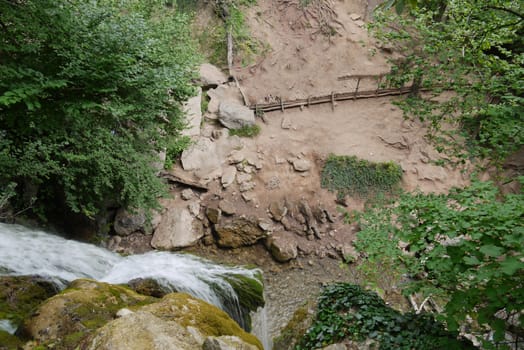 A top-down view of a waterfall flowing over rocks. Forming below a small pond and running further into the depths of the forest by a transparent stream.