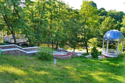 Beautiful nature in the park on a clear sunny afternoon. With a green slope and a descending downhill leading to the gazebo with a domed roof.