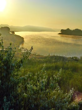 A beautiful picture of the sunrise over the river with picturesque shores with green grass and trees wrapped in morning mist.