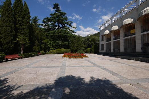 A beautiful flowerbed with bright flowers in the middle of the paved area with standing benches on the edges for rest and designer bushes of greenery.