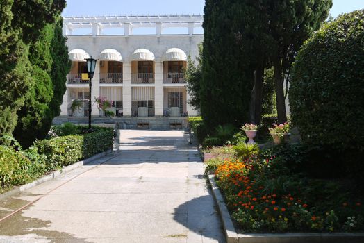 A stone alley with beautiful decorative flower beds and trees on the sides, leading to the building's hull