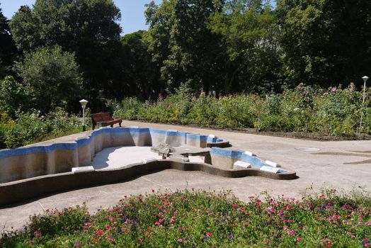 An empty pool in the middle of a park with crooked benches and large flower beds