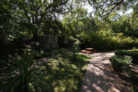 Small wooden benches with a table standing in the shade of trees in a cozy place in the park next to the path and a large stone lying near the bushes.
