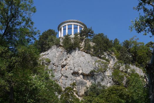 on the top of a rocky hill is an architectural construction of a circular shape with columns of white against the background of the celestial blue