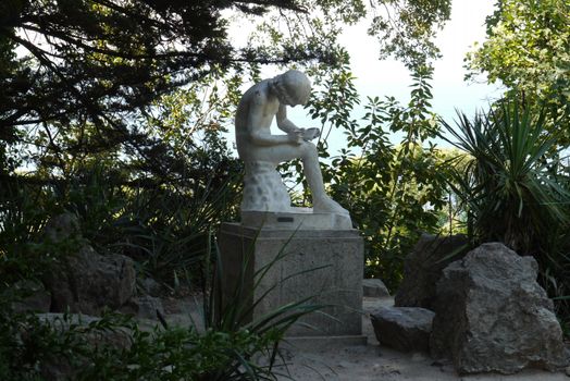 A gypsum white color statue in the shade of a tree among the bushes. It depicts a person writing something to a notebook.