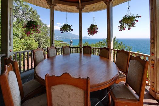 A massive round wooden table with chairs standing in a gazebo for relaxing with hanging flowerpots with flowers and a magnificent view of the sea and mountain slopes in the distance.