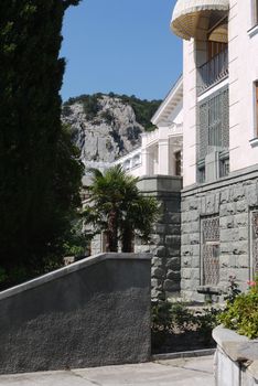 Beautiful architecture of the building and the courtyard with the growing palm trees nearby and the stony slope of a small mountain behind it.