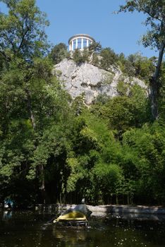 The arbor on top of a rocky cliff with growing green trees on the shore of a pond located in the park.