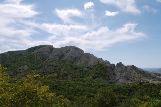 The rocky mountain peak against the background of a bright sky with transparent clouds overgrown on the slopes by a rare and below a dense green forest.