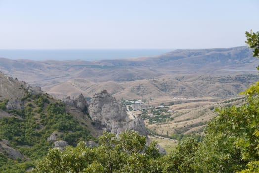 The houses of a small village lying in a valley among the hills without vegetation. With the blue sea stretching out beyond the horizon in the distance.