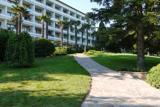 Alley is lined with stone in the middle of a green cropped lawn in an exotic park in front of a snow-white tourist hotel.