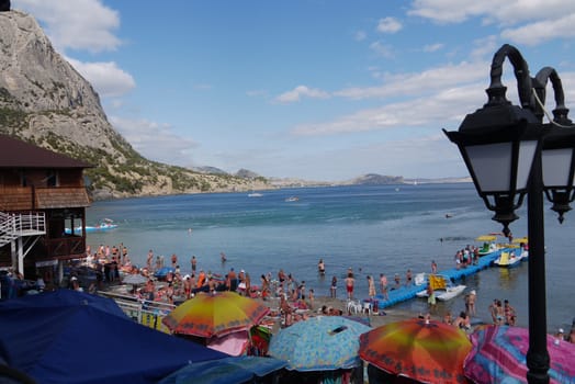A beach full of tourists and colorful umbrellas by the sea coast in the background of a high rocky mountain
