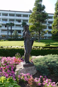 Sculpture of a woman on a green lawn standing on a stone with an inscription on the plinth of Flora with growing flowers beside the hotel with balconies.