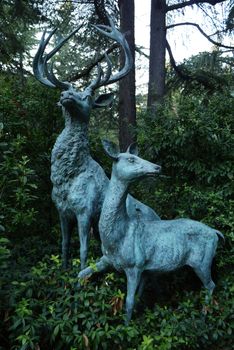 Sculptures of a deer with large spreading horns and deer anxiously looking to the side in the park among the dense green thickets of bushes.