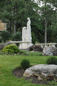 A tall sculpture standing on a white pedestal in a park next to tall pine trees on one side and a green lawn with flowers on the other.