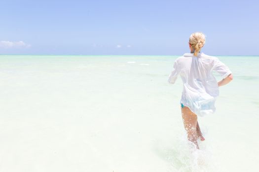Young slim fit woman wearing white beach tunic running in sea water making water splashes with her legs. Vacation concept. Summer mood. Tropical beach setting. Paje, Zanzibar, Tanzania.