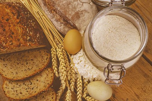 Loafs of bread, slices of bread, wheat flour, eggs and ears of grain on wood background. Rustic and rural concept. Close up. Flat lay.  