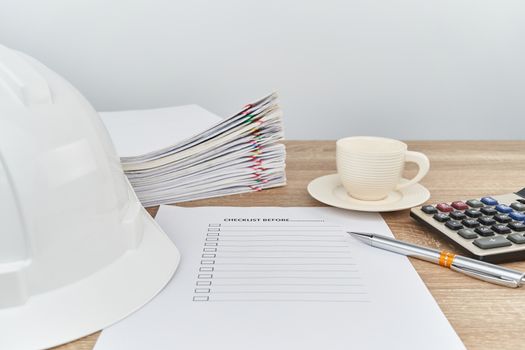Pen on checklist with calculator have blur white engineer hat and cup of coffee with pile overload document of report and receipt on wooden table with white background and copy space.