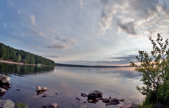 the evening sky over the lake, silhouettes of clouds illuminated by the sun, forest-covered, rocky shore of the lake