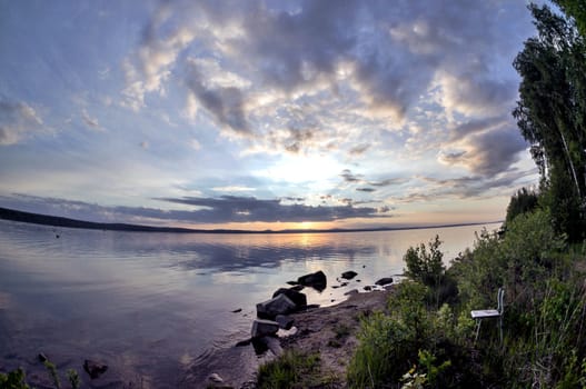 the evening sky over the lake, silhouettes of clouds illuminated by the sun, forest-covered, rocky shore of the lake