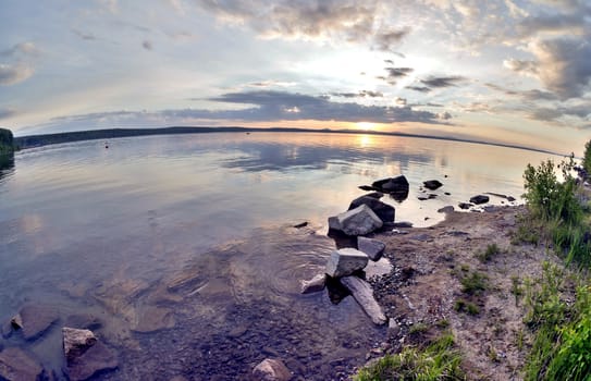 the evening sky over the lake, silhouettes of clouds illuminated by the sun, forest-covered, rocky shore of the lake
