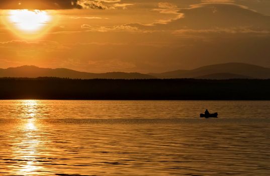 orange sunset over the lake, the sun lights up the clouds, a lone fisherman on a boat