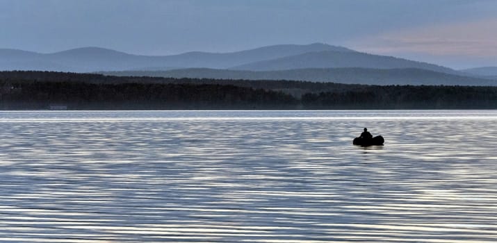 evening gray-blue lake and sky, a lone fisherman on a boat, in the distance you can see the silhouettes of the Ural mountains, the southern Urals, lake Uvildy