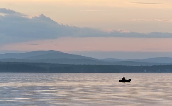 evening gray-blue lake and sky, a lone fisherman on a boat, in the distance you can see the silhouettes of the Ural mountains, the southern Urals, lake Uvildy