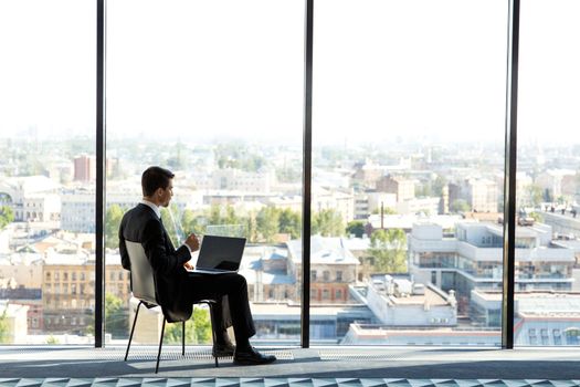 Business man sitting in modern office with panoramic windows, typing on his laptop and srinking coffee