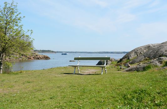 Empty green and white bench on a grassy hilltop above the water on Suomenlinna, a ferry sails past in the background