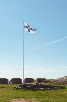 Finland state flag flying high on a flagpole on Suomenlinna sea fortress. A row of disused cannons lies below on the grass.