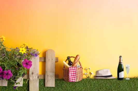 Summer picnic basket with bread, cheese and wine on grass with garden fence and flowers