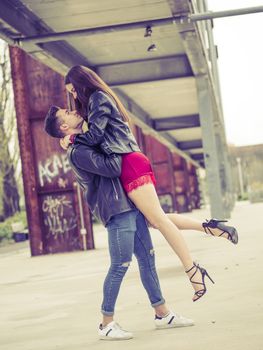 Side view of trendy man holding stylish girl in dress and heels above ground standing on grungy street background.